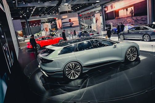 A slick, modern exhibition space featuring several high-end cars. A white sedan is prominently displayed on a circular platform, surrounded by other luxury vehicles in various colors, including a striking red car. The environment is bustling with people, likely attendees and exhibitors, moving around the cars. Large digital screens and branded banners adorn the background.