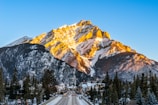 A majestic mountain dominates the landscape, partially covered with snow and bathed in warm sunlight. Below, a quaint town with houses lines the street leading towards the mountain, flanked by tall evergreen trees. The bright blue sky creates a vivid backdrop.