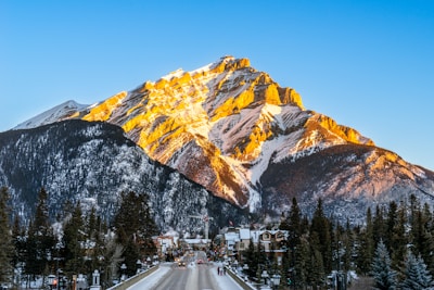 A majestic mountain dominates the landscape, partially covered with snow and bathed in warm sunlight. Below, a quaint town with houses lines the street leading towards the mountain, flanked by tall evergreen trees. The bright blue sky creates a vivid backdrop.