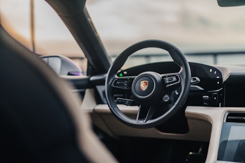 A luxurious car interior featuring a sleek, modern dashboard with a digital display and a prominent steering wheel bearing the Porsche logo. The design boasts premium materials and a sophisticated aesthetic.