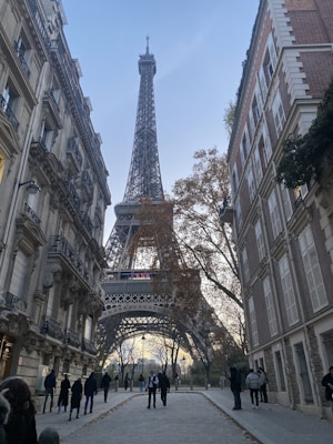 A street view leading towards the Eiffel Tower, framed by classic Parisian buildings on either side. People walk along the cobblestone path beneath the leafless trees, creating a serene urban scene.