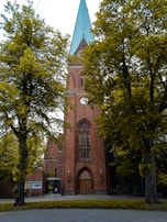 A tall, brick church with a pointed green roof is flanked by large, leafy trees. The church features a large arched wooden door and a tall, narrow stained glass window above. Two lamps are located on either side of the entrance.