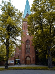 A tall, brick church with a pointed green roof is flanked by large, leafy trees. The church features a large arched wooden door and a tall, narrow stained glass window above. Two lamps are located on either side of the entrance.