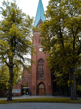 A tall, brick church with a pointed green roof is flanked by large, leafy trees. The church features a large arched wooden door and a tall, narrow stained glass window above. Two lamps are located on either side of the entrance.