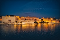 A beautifully lit cityscape reflecting on a tranquil body of water during twilight. The architecture features grand buildings with intricate details, creating a majestic atmosphere under the deep blue evening sky.