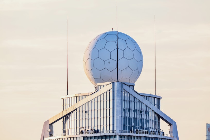 A modern building features a geodesic dome with hexagonal patterns at the top, likely housing a radar or communication system. The structure has multiple tall antennas extending upwards and large glass windows reflecting the soft, pastel colors of the sky.