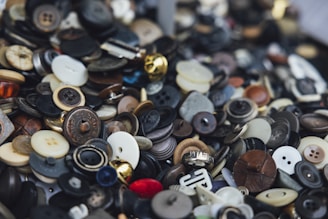 A large collection of assorted buttons in various colors, sizes, and materials. The buttons are piled together, creating a chaotic and dense visual appearance. Some buttons are shiny, metallic, while others are matte and made of plastic or wood. There are both old, worn-out buttons and new, pristine ones.