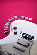 An electric guitar partially visible against a vibrant red background, with multiple colorful guitar picks arranged in an arc nearby. The guitar features cream-colored knobs and a set of strings leading to a black bridge.