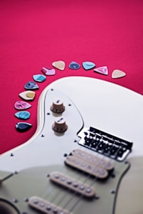 An electric guitar partially visible against a vibrant red background, with multiple colorful guitar picks arranged in an arc nearby. The guitar features cream-colored knobs and a set of strings leading to a black bridge.