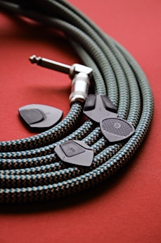 A coiled, textured guitar cable with a checkerboard pattern in green and beige lies on a red surface. Beside the cable, four black guitar picks with varying textures and markings are scattered.