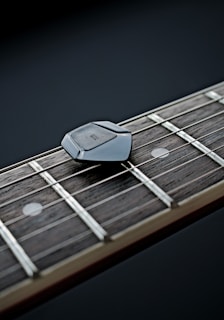 A close-up view of a guitar neck, focusing on the strings and frets. There is a black guitar pick resting on the strings.