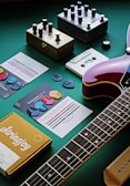 A collection of guitar-related items is arranged on a green surface. Several guitar effect pedals with various knobs are placed beside music cards featuring colorful guitar picks. A guitar is partially visible, focusing on the neck and body. A yellow box labeled 'Stringjoy' appears to contain guitar strings.