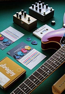 A collection of guitar-related items is arranged on a green surface. Several guitar effect pedals with various knobs are placed beside music cards featuring colorful guitar picks. A guitar is partially visible, focusing on the neck and body. A yellow box labeled 'Stringjoy' appears to contain guitar strings.