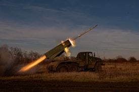A military vehicle is positioned in an open field, launching a missile into the sky. The missile is in mid-air, with bright flames and smoke trailing from its base, indicating active launch. The vehicle is painted with camouflage colors, blending with the surrounding dry grass and leafless trees.