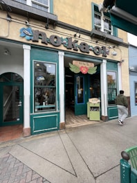 The image features the exterior entrance of a small shop or cafe with teal and white framing. The sign above the entrance reads 'No Ka Oi' decorated with floral motifs. There is a sidewalk in front, and a person is seen walking towards the entrance.