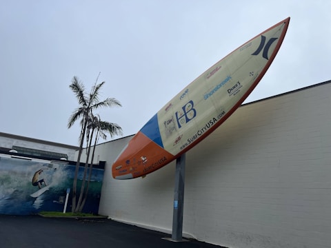 A large surfboard is mounted upright against a building. The surfboard features various logos and the text 'SURFCITYUSA.com'. A mural painting of a surfer riding a wave is visible on the building wall. Tall palm trees are situated nearby, under a cloudy sky.