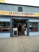 A restaurant and pub with a sign reading 'The Longboard' above the entrance. The exterior features blue paneling and large windows displaying various items inside. There's a wooden door, open to welcome guests, and a chalkboard sign outside on the sidewalk.