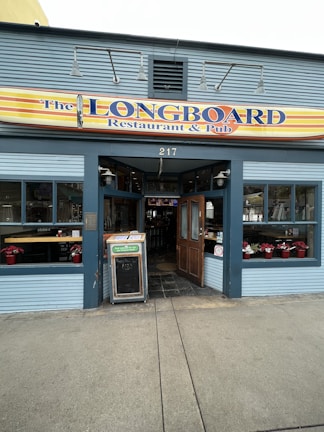 A restaurant and pub with a sign reading 'The Longboard' above the entrance. The exterior features blue paneling and large windows displaying various items inside. There's a wooden door, open to welcome guests, and a chalkboard sign outside on the sidewalk.