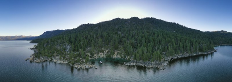 A large forested island surrounded by a calm body of water under a clear blue sky. The island is densely covered with green coniferous trees and has rocky edges. The tranquil water reflects the hues of the surrounding landscape.