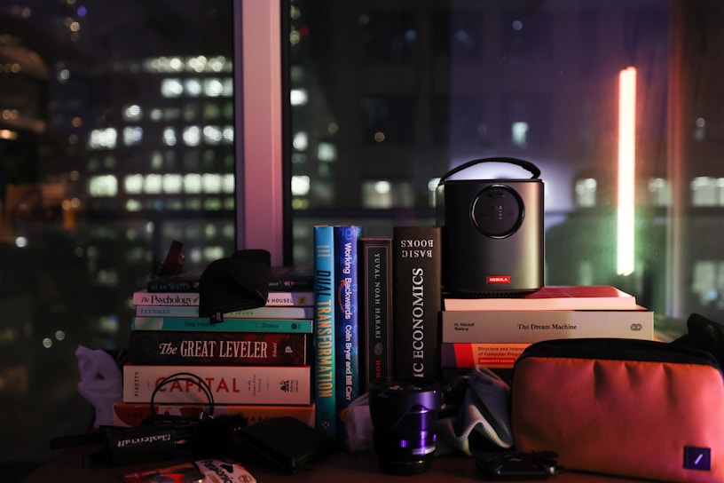 A collection of books stacked on a table is visible, with titles such as 'The Great Leveler' and 'Basic Economics.' A small projector is placed on top of some books, and there are various other items like a camera lens and a pouch. Through the window in the background, the cityscape featuring blurred lights of buildings suggests a nighttime setting.