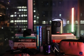 A collection of books stacked on a table is visible, with titles such as 'The Great Leveler' and 'Basic Economics.' A small projector is placed on top of some books, and there are various other items like a camera lens and a pouch. Through the window in the background, the cityscape featuring blurred lights of buildings suggests a nighttime setting.