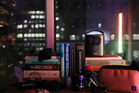 A collection of books stacked on a table is visible, with titles such as 'The Great Leveler' and 'Basic Economics.' A small projector is placed on top of some books, and there are various other items like a camera lens and a pouch. Through the window in the background, the cityscape featuring blurred lights of buildings suggests a nighttime setting.