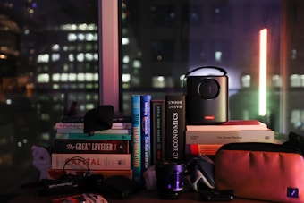 A collection of books stacked on a table is visible, with titles such as 'The Great Leveler' and 'Basic Economics.' A small projector is placed on top of some books, and there are various other items like a camera lens and a pouch. Through the window in the background, the cityscape featuring blurred lights of buildings suggests a nighttime setting.