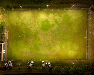 Aerial view of a brightly lit rectangular soccer field with two goalposts at either end. The surrounding area includes a boundary fence, several parked cars on one side, and a small building adjacent to the field. The grass has varying shades of green with some patchy areas.