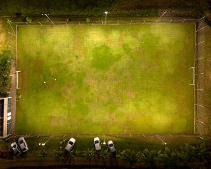 Aerial view of a brightly lit rectangular soccer field with two goalposts at either end. The surrounding area includes a boundary fence, several parked cars on one side, and a small building adjacent to the field. The grass has varying shades of green with some patchy areas.