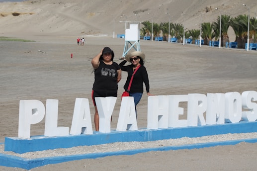 Two people standing next to a large white sign that reads 'Playa Hermosa' on a sandy beach. The background features a series of palm trees along with some distant beachgoers and structures. The individuals are dressed casually, one wearing a hat.