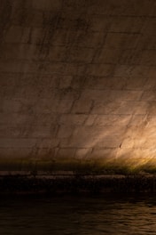 A textured concrete wall is illuminated by a soft light, creating a warm glow. The surface shows signs of age and wear, with visible moss or algae growth near the waterline. The bottom of the wall meets a calm body of water that reflects the light.