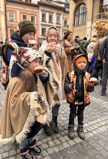 A group of children dressed in traditional, colorful clothing are standing on a cobblestone street, eating pastries. They appear to be part of a festive gathering or celebration. In the background, there is a crowd of people and buildings with ornate architecture.