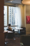 A person sits alone at a round table in a softly lit room, looking at a phone. Large windows reveal tall buildings outside. Sheer white curtains partly cover the windows, and the interior features muted tones and low lighting, suggesting a quiet, contemplative atmosphere.
