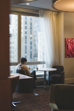A person sits alone at a round table in a softly lit room, looking at a phone. Large windows reveal tall buildings outside. Sheer white curtains partly cover the windows, and the interior features muted tones and low lighting, suggesting a quiet, contemplative atmosphere.