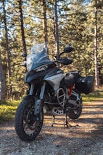 A motorcycle stands on a dirt path in a wooded area. The bike is equipped with off-road tires and a windshield, showcasing a rugged and adventurous design. Sunlight filters through the tall trees, casting shadows on the ground and illuminating the motorcycle.