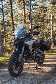 A motorcycle stands on a dirt path in a wooded area. The bike is equipped with off-road tires and a windshield, showcasing a rugged and adventurous design. Sunlight filters through the tall trees, casting shadows on the ground and illuminating the motorcycle.