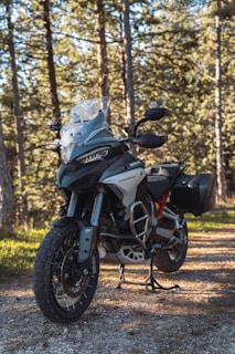 A motorcycle stands on a dirt path in a wooded area. The bike is equipped with off-road tires and a windshield, showcasing a rugged and adventurous design. Sunlight filters through the tall trees, casting shadows on the ground and illuminating the motorcycle.
