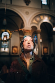 A person is looking upwards with a contemplative expression, standing in an ornate, dimly-lit interior space that appears to be a church. The architecture is highlighted by arches, columns, and stained glass windows, with warm, ambient light casting a serene glow.