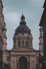 A large, ornate building with classical architecture, featuring a prominent, dark-colored dome. The facade is adorned with columns and detailed sculptures, including a depiction of religious scenes above the entrance. The inscription 'EGO SUM VIA VERITAS ET VITA' is visible across the archway.
