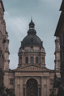A large, ornate building with classical architecture, featuring a prominent, dark-colored dome. The facade is adorned with columns and detailed sculptures, including a depiction of religious scenes above the entrance. The inscription 'EGO SUM VIA VERITAS ET VITA' is visible across the archway.