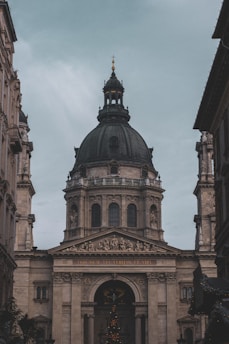A large, ornate building with classical architecture, featuring a prominent, dark-colored dome. The facade is adorned with columns and detailed sculptures, including a depiction of religious scenes above the entrance. The inscription 'EGO SUM VIA VERITAS ET VITA' is visible across the archway.
