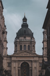 A large, ornate building with classical architecture, featuring a prominent, dark-colored dome. The facade is adorned with columns and detailed sculptures, including a depiction of religious scenes above the entrance. The inscription 'EGO SUM VIA VERITAS ET VITA' is visible across the archway.