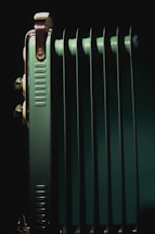 A close-up view of a metallic, green radiator with multiple vertical fins. The lighting highlights the metallic components and edges, creating a contrast with the dark background.