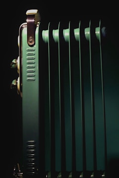 A close-up view of a metallic, green radiator with multiple vertical fins. The lighting highlights the metallic components and edges, creating a contrast with the dark background.