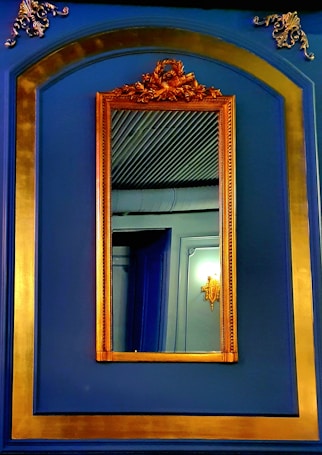 An ornate mirror with a golden frame is mounted on a deep blue wall adorned with gold trim. The interior reflection shows intricate wall molding and a sconce light fixture, creating a luxurious and elegant atmosphere.