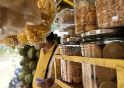 A market or street vendor stall displaying various snacks in transparent containers. There are glass jars filled with different types of snacks, including what appears to be chips and pastries. The setting has a rustic feel with a blurred background showing a person in a yellow shirt.