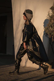 A person walking outdoors wearing a dark leather outfit, including a long coat and pants. They also have a brown and gray fur hat. The background features a textured white wall and a leafy plant casting shadows.