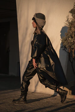 A person walking outdoors wearing a dark leather outfit, including a long coat and pants. They also have a brown and gray fur hat. The background features a textured white wall and a leafy plant casting shadows.