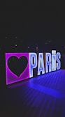 A glowing sign with bold letters spelling 'PARIS,' accompanied by a large heart shape to the left. The sign is illuminated with neon lights, giving off vibrant purple and blue hues. It appears to be displayed outdoors, possibly at night, against a dark background.