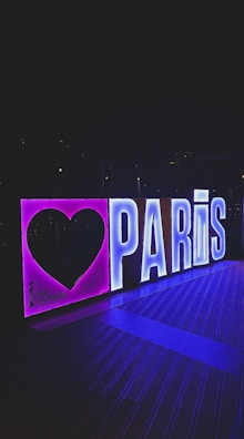 A glowing sign with bold letters spelling 'PARIS,' accompanied by a large heart shape to the left. The sign is illuminated with neon lights, giving off vibrant purple and blue hues. It appears to be displayed outdoors, possibly at night, against a dark background.